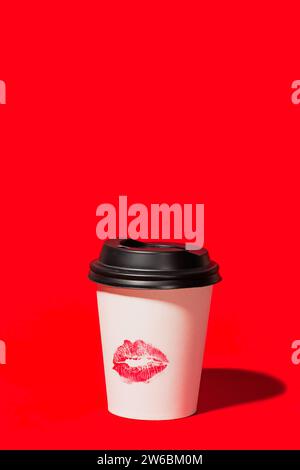 Eine einzelne weiße Einwegkaffeetasse mit schwarzem Deckel und rotem Lippenstift-Kiss steht auf einem leuchtend roten Hintergrund und sorgt für einen auffälligen und romantischen Ausdruck Stockfoto