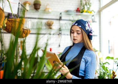 Floristin im Kopftuch mit Tablette mit Checkliste während der Arbeit im Blumenladen bei der Inventur von Topfpflanzen Stockfoto