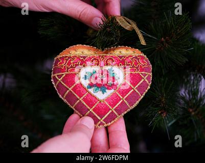 Eine Frau, die ein rotes Kreuzstich mit zeitloser Eleganz an einen Weihnachtsbaum hängt. Dieses herzförmige Weihnachtsschmuck mit roten Blumen e Stockfoto