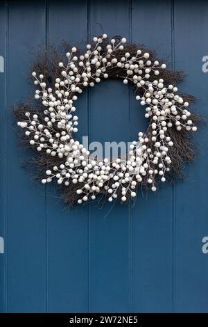 Nahaufnahme eines wunderschönen weißen Weihnachtskranzes, der an einer blauen Holztür im Dorf Lacock, Wiltshire, England, Großbritannien hängt. Dezember Stockfoto