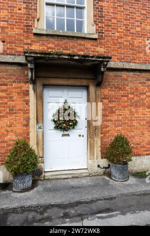 Nahaufnahme einer weißen Eingangstür mit Weihnachtskranz auf einem roten gemauerten Haus im National Trust Dorf Lacock, Wiltshire, England, Großbritannien Stockfoto