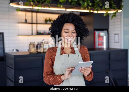 Junge afroamerikanische Frau mit Schürze, die in einem Restaurant und einem Esszimmer arbeitet, lächelnd steht und ein Tablet benutzt. Stockfoto