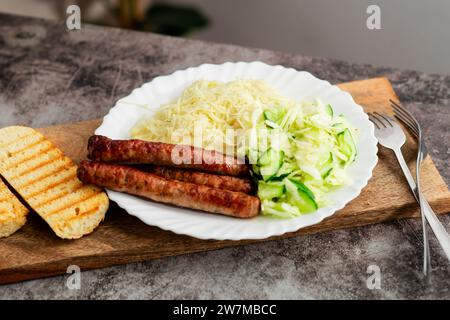 Traditionelle Küche: Würstchen, Kartoffelpüree und ein würziger Kohl-Gurkensalat auf einem weißen Teller, auf einem rustikalen Holztisch Stockfoto