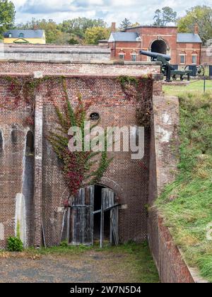 Die verwitterte hölzerne Kellertür eines roten Ziegelbaus, geschmückt mit Kletterstöcken, mit einer Kanone als Hintergrund in Fort Washington, Virginia. Stockfoto