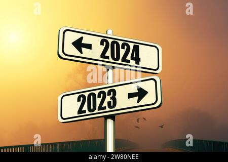 Straßenschilder zeigen den Weg zum Neujahr 2024 und das Alte Jahr 2023 auf Einem blauen Himmel mit Nebel und Sonnenuntergang, kreative Idee. Wählen Sie Eine neue Straße, ein Konzept. Stockfoto