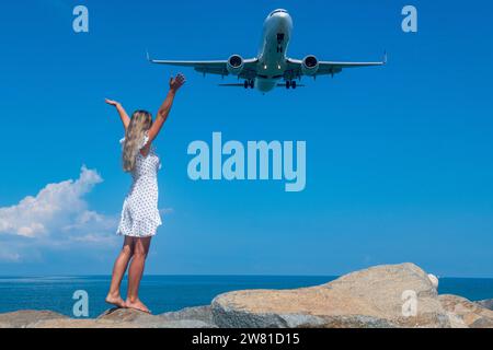 Ozeanische Verbindung: Ein Mädchen in einem weißen Kleid auf Steinen trifft auf ein fliegendes Flugzeug Stockfoto