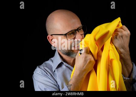 Kahler, bärtiger Mann mit einer Brille, der ein gelbes Tuch über dem Kopf vor schwarzem Hintergrund hält. Stockfoto