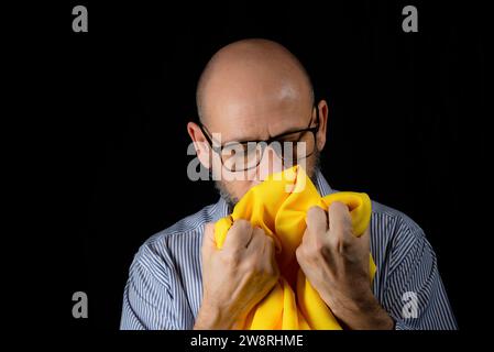 Kahler, bärtiger Mann mit einer Brille, der ein gelbes Tuch über dem Kopf vor schwarzem Hintergrund hält. Stockfoto