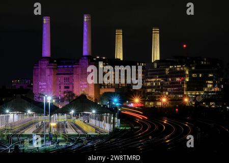 London, Großbritannien. Dezember 2023. Pendlerzüge, die auf einem langen Lichtweg zu sehen sind, fahren an der Battersea Power Station vorbei, die in der Abenddämmerung in einem festlichen Design des Künstlers David Hockney beleuchtet wird. Quelle: Eleventh Photography/Alamy Live News Stockfoto