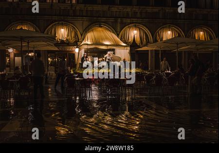 Markusplatz in Venedig, Italien, am regnerischen Abend mit einer Gruppe von Musikern, die unter der Markise des Restaurants spielen; Lichter reflektieren in Pfützen Stockfoto