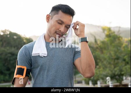 Ein müder asiatischer Mann in Sportbekleidung wischt sich beim Laufen im Freien den Schweiß über sein Gesicht. Intensives Workout, erschöpft Stockfoto