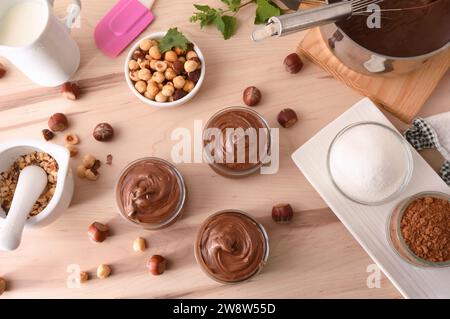 Hausgemachte Haselnusscreme in Schüsseln mit Nüssen, Schokolade, anderen Zutaten und Werkzeugen auf Holzbank. Draufsicht. Stockfoto