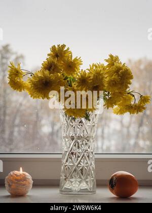 Senkrechter Blick auf gelbe Chrysanthemen in einer Glasvase, Kerzenlicht und ein leuchtendes orangenes Persimmon auf einem weißen mit einem winterlichen Waldpanorama. Stockfoto