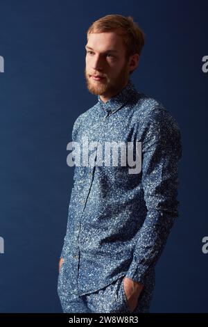 Männer-, Mode- und schicke Kleidung im Studio mit einzigartigem Stil, Denim und trendy auf blauem Hintergrund. Irischer Odel mit Hipster-Bart, rotes Haar mit Serious Stockfoto