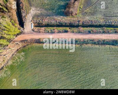 Von einem erhöhten Aussichtspunkt aus zeigt das Bild eine ruhige Landschaft mit einem separaten Pfad, der durch die Landschaft führt. Der Weg dient als Trennlinie zwischen einem strukturierten grünen Feld auf der einen Seite und einem dunkleren, möglicherweise bewaldeten Gebiet auf der anderen Seite. Die Schatten sind kurz, was darauf hindeutet, dass ein Foto kurz vor Mittag aufgenommen wurde. Die Felder zeigen Veränderungen im Pflanzenwachstum oder der Graslänge, wodurch Muster entstehen, die durch das Sonnenlicht hervorgehoben werden. Luftperspektive von Landweg und Feldern. Hochwertige Fotos Stockfoto