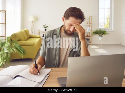 Müder Mann, der an Kopfschmerzen leidet und an seinem Arbeitsplatz mit einem Laptop arbeitet. Stockfoto