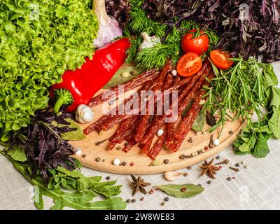 Dünne geräucherte Würstchen mit frischen Kräutern, Paprika, Tomaten und anderen Gewürzen auf einem Schneidebrett Stockfoto