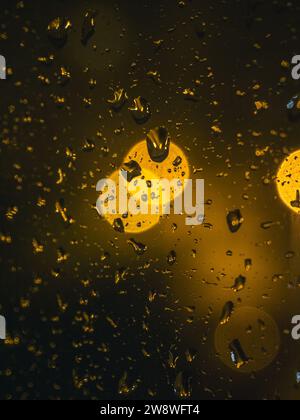 Regentropfen an einem Fenster während der Nacht mit bunten, verschwommenen Stadtlichtern, Straßenlaternen an der Außenseite, warmen Farben, stimmungsvoller Atmosphäre Stockfoto