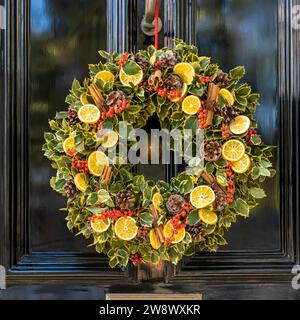 Ein Adventskranz aus stechpalmenzweigen, Tannenzapfen, roten Beeren, getrockneten Orangen und Zimtstangen, an einer grauen Londoner Tür. Quadratischer Rahmen. Stockfoto