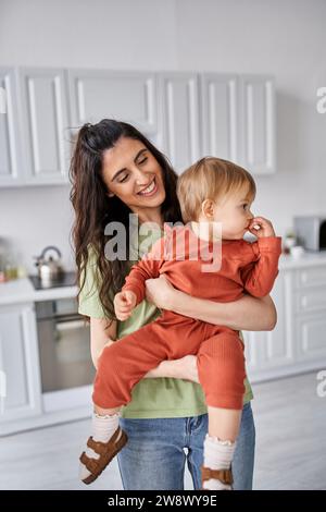 Fröhliche brünette Frau gewinnt Haushaltswaren, hält ihr Baby und lächelt glücklich, Familienkonzept Stockfoto