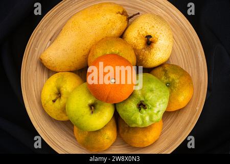 Mit Blick von oben auf einen Haufen reifer frischer Guave-Früchte, rote Tomaten, Orangen, Birnen und Äpfel in Bambusplatte auf schwarzem Hintergrund. Stockfoto