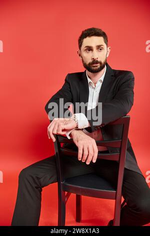 Charismatischer bärtiger Geschäftsmann in schwarzem Anzug, der auf einem Stuhl sitzt und vor rotem Hintergrund wegblickt Stockfoto