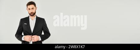 Charismatischer bärtiger Geschäftsmann, der schwarzen Anzug zuknöpft und die Kamera auf grauem Banner ansieht Stockfoto
