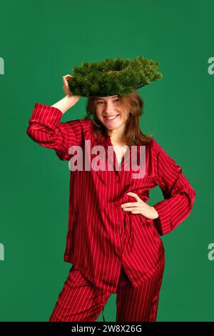 Schönes junges Mädchen im schönen roten Seidenschlafanzug posiert mit dem Neujahrskranz, der ihn auf ihrem Kopf vor grünem Hintergrund hält. Konzept von Stockfoto