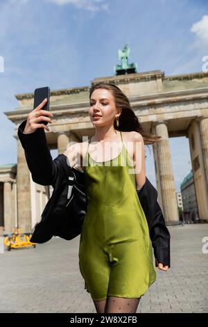 Modische Frau in Jacke und Seidenkleid beim Selfie am Brandenburger Tor in Berlin Stockfoto