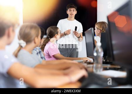 Ein freundlicher junger Lehrer steht neben einem interaktiven Whiteboard und erzählt Jugendlichen im Computerunterricht neues Material Stockfoto