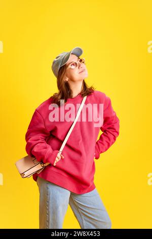 Porträt in voller Länge eines glücklichen, fröhlichen Studenten, übergroße Kleidung und trendige Sonnenbrille vor lebhaftem gelbem Hintergrund. Stockfoto