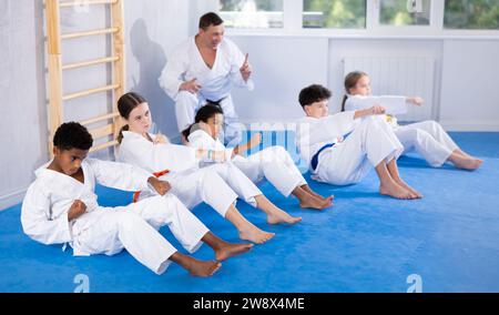 Gruppe multinationaler Kinder im Kimono trainieren Bauchmuskeln und machen gleichzeitig Schläge während Karate- oder Judo-Trainings Stockfoto
