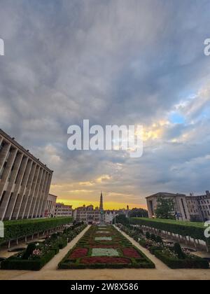 Der Mont des Arts bietet eine atemberaubende Aussicht auf Brüssel mit einem dramatischen und dynamischen Himmel über dem Kopf. Die Wolken teilen sich leicht und zeigen einen Hauch von Blau und das sanfte Leuchten der untergehenden Sonne, die ein goldenes Licht auf die Stadt darunter wirft. Dieser Ort ist ein kulturelles Zentrum, mit dem Turm des Rathauses in der Ferne, was die historische Bedeutung der Szene unterstreicht. Der gepflegte Garten im Vordergrund mit seinen geometrischen Mustern und dem lebhaften Grün steht im Kontrast zur urbanen Architektur und schafft eine Mischung aus Natur und Zivilisation. Dramatischer Himmel über dem Mont des Arts. Hochwertige Fotos Stockfoto