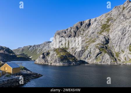 Nusfjord, Lofoten: Ein ruhiger Fjord, flankiert von steilen Klippen mit einer traditionellen gelben Fischerhütte, die Norwegens raue Küstenschönheit widerspiegelt Stockfoto