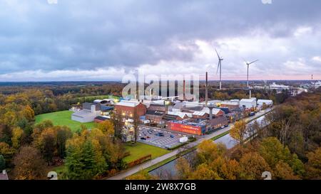 Dieses Luftbild bietet eine überzeugende Gegenüberstellung von alter und neuer Industrie mit einem weitläufigen Blick auf einen traditionellen Fabrikkomplex vor der Moderne hoch aufragender Windturbinen. Eingebettet in eine grüne Waldlandschaft, gibt die Industrieanlage mit ihren hohen Schornsteinen Rauchwolken ab, während die sauberen Energieerzeuger still im Hintergrund stehen und einen Übergang zu nachhaltigen Praktiken symbolisieren. Der bewölkte Himmel und das Licht der Morgen- oder Abenddämmerung deuten auf eine Zeit des Wandels hin, nicht nur des Tages, sondern vielleicht auch einer Ära der Öko-industriellen Evolution: Windturbinen mit Blick auf einen Stockfoto