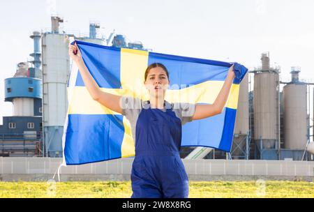 Auf dem Land steht ein frustrierter Arbeiter im Overall hinter dem Fabrikgebäude mit der Flagge Schwedens Stockfoto