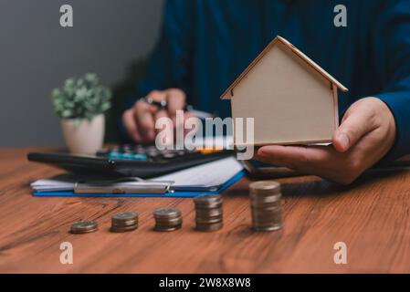 Drücken Sie den Taschenrechner von Hand, halten Sie ein Modellhaus und stapeln Sie Münzen auf den Tisch. Stockfoto