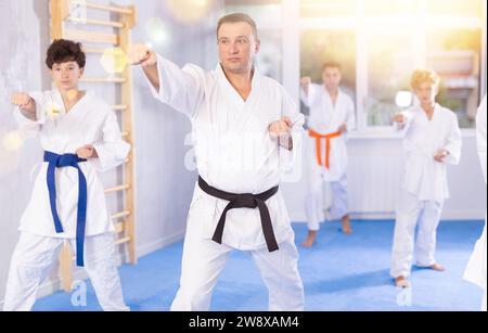 Karatekinder im Kimono führen während des Gruppentrainings Kata-Moves mit ihrem Lehrer im Vordergrund im Fitnessstudio durch Stockfoto