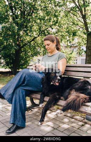 Reife Frau mit australischem Schäferhund mit Smartphone auf der Bank im Park Stockfoto