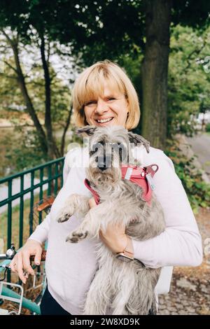 Porträt einer lächelnden blonden Frau, die einen süßen Hund im öffentlichen Park hält Stockfoto