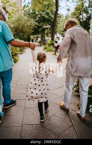 Großeltern halten Hände von Enkelin, während sie auf einem Fußweg an der Straße laufen Stockfoto