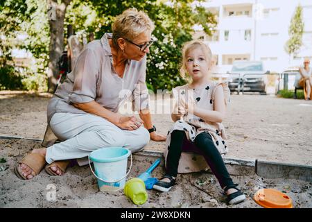 Großmutter spricht mit Enkelin, die im Sandkasten sitzt, während sie im Park spielt Stockfoto