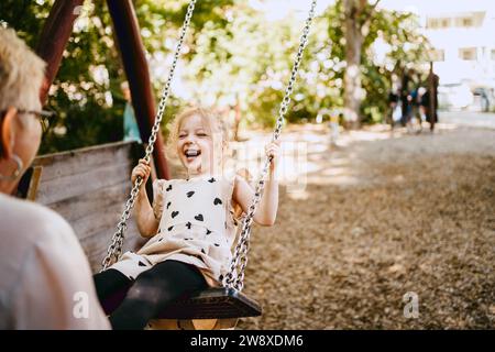 Glückliches Mädchen lacht, während es mit Großmutter im Park auf der Schaukel spielt Stockfoto