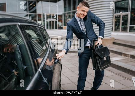 Geschäftsmann, der mit dem Smartphone spricht, während er die Autotür in der Nähe des Bürogebäudes öffnet Stockfoto