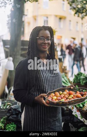 Porträt einer lächelnden Verkäuferin, die ein Tablett mit frischen Pflaumen hält, während sie auf dem Bauernmarkt steht Stockfoto