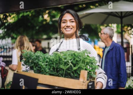 Junge lächelnde Floristin, die eine Pflanzenkiste auf dem Flohmarkt hält Stockfoto