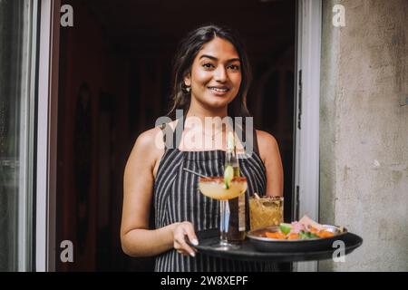 Porträt einer lächelnden Kellnerin, die ein Tablett mit Speisen und Getränken am Eingang der Bar hält Stockfoto