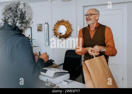 Lächelnder, männlicher Unternehmer, der weiblichen Kundin Papiertüte gibt, während sie im Geschäft einkaufen Stockfoto