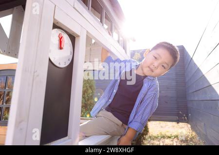 Ein asiatischer kleiner Junge schaut aus einem Holzhaus. Ein koreanisches Kind spielt im Garten. Niedliches Kind in lässiger Kleidung draußen auf einem sonnigen Stockfoto