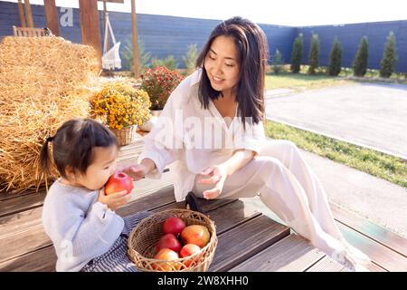 Asiatische attraktive Mutter und ihre süße Tochter im Hinterhof des Hauses. Ein kleines süßes Mädchen isst draußen Äpfel. Eine charmante Koreanerin gibt Stockfoto